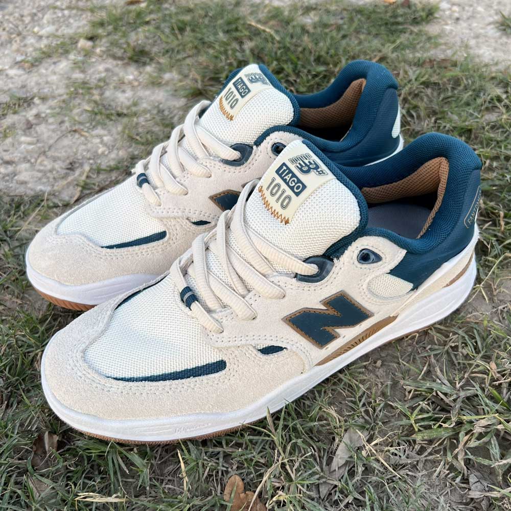 A pair of 1010 BL Tiago Lemos Beige Blue Suede New Balance Numeric Skateboarding Shoes rest on grass and dirt, showcasing suede and mesh details with branded labels—ideal for any skateboarding session.
