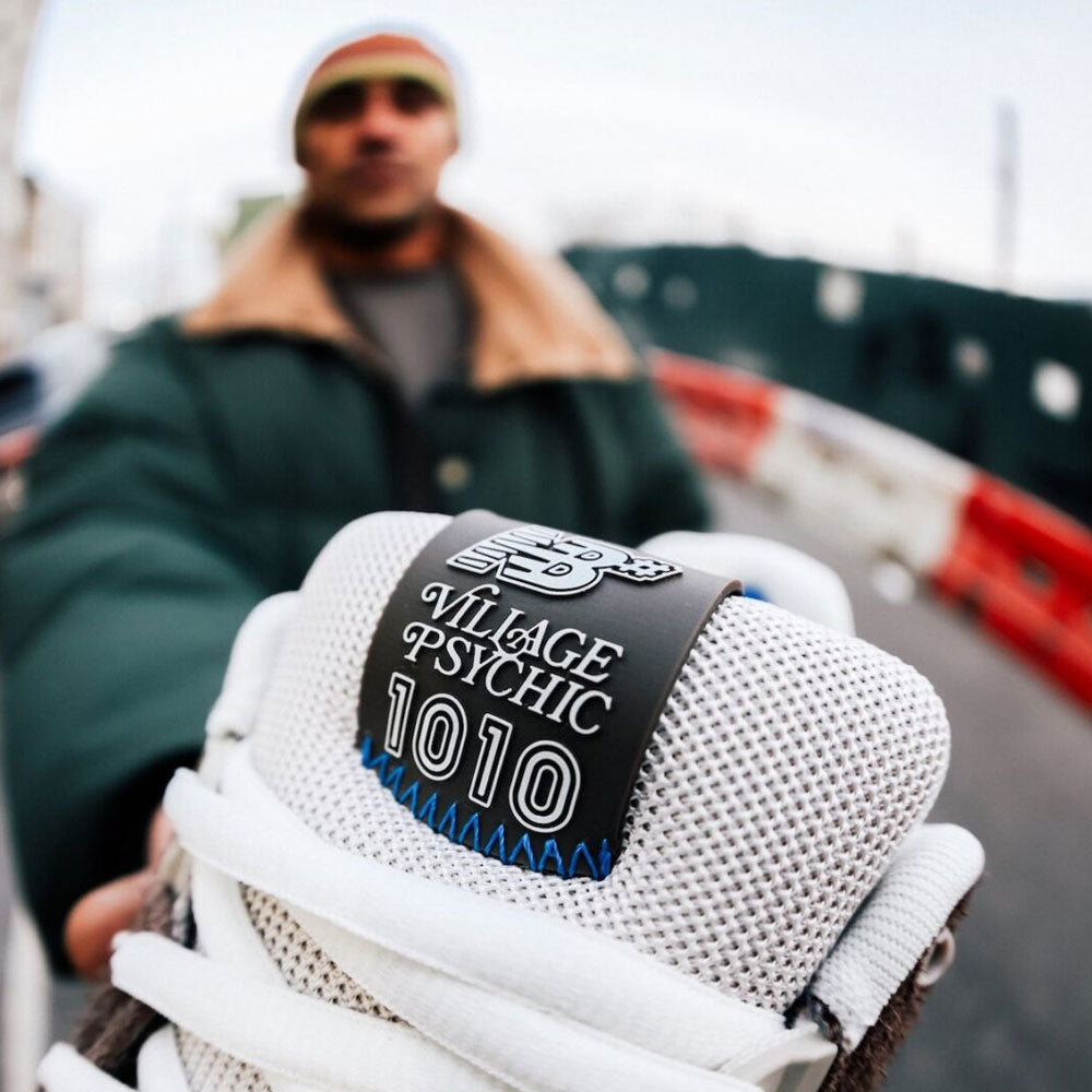 A person in a green coat holds a 1010 SV Village Psychic Tiago Lemos Gray Blue Suede New Balance Numeric Skateboarding Shoe close to the camera, "VILLAGE PSYCHIC 1010" visible on the tongue, against a blurred urban street backdrop.