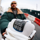 A person in a green coat holds a 1010 SV Village Psychic Tiago Lemos Gray Blue Suede New Balance Numeric Skateboarding Shoe close to the camera, "VILLAGE PSYCHIC 1010" visible on the tongue, against a blurred urban street backdrop.