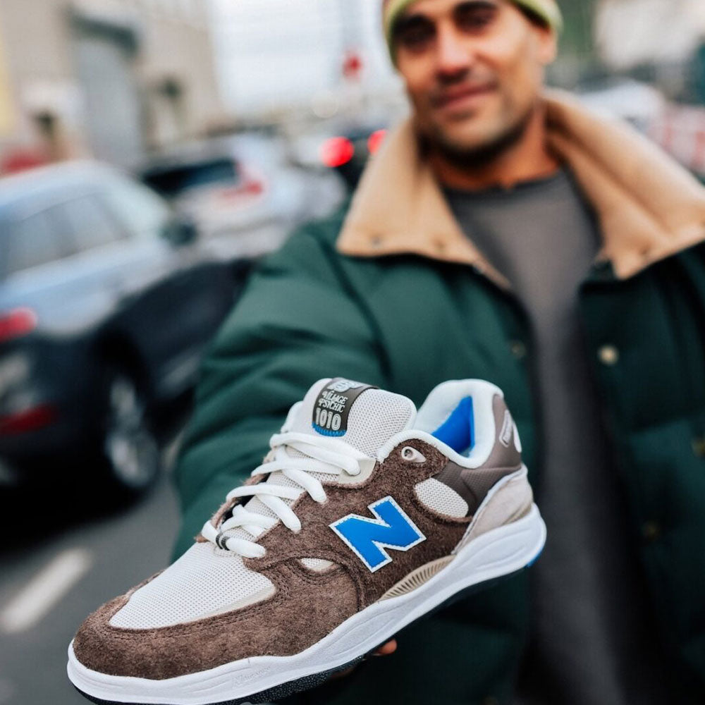 A person in a green jacket with a beige collar holds out the gray and blue suede New Balance Numeric 1010 SV Village Psychic Tiago Lemos skateboarding shoe, highlighting its comfort as the city street blurs in the background.