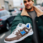 A person in a green jacket with a beige collar holds out the gray and blue suede New Balance Numeric 1010 SV Village Psychic Tiago Lemos skateboarding shoe, highlighting its comfort as the city street blurs in the background.