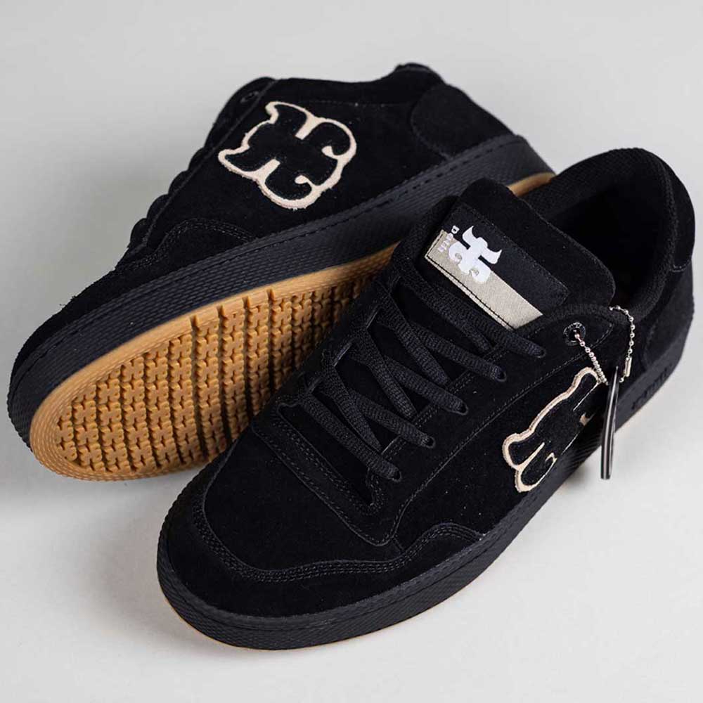 The 1985 Black Tan I Path Shoes Skateboarding feature black suede uppers, gum cupsoles, black laces, and a white embroidered abstract logo on the sides and tongue. One shoe stands upright; the other lies flat, showing the sole’s texture.