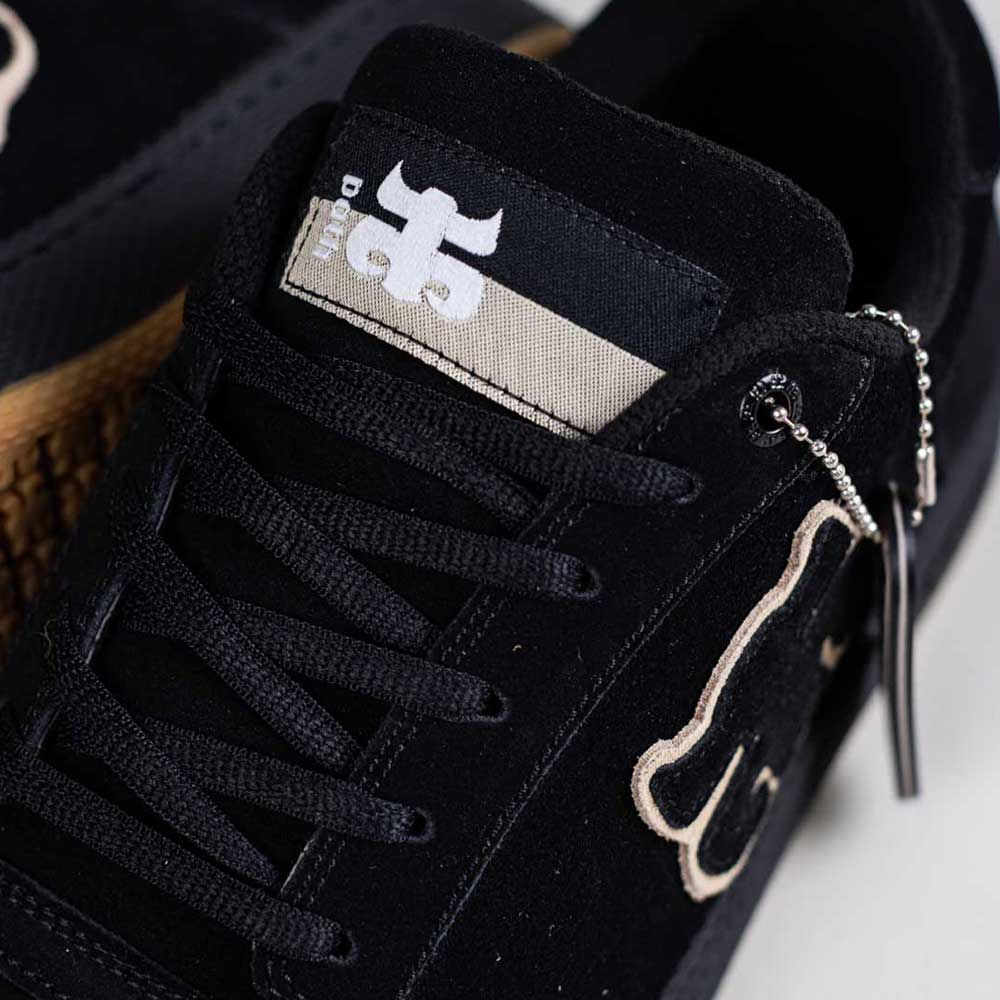 Close-up of the 1985 Black Tan I Path Shoes Skateboarding, featuring black suede, black laces, cupsole construction, side silver zipper pull, white embroidered bone design, and a black and white tongue label with a stylized logo.