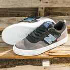 A pair of 306 TRI New Balance Numeric Jamie Foy Suede Shoes in grey and black with white soles and blue N logos, one upright and one on its side to show the sole, displayed on a wooden surface.