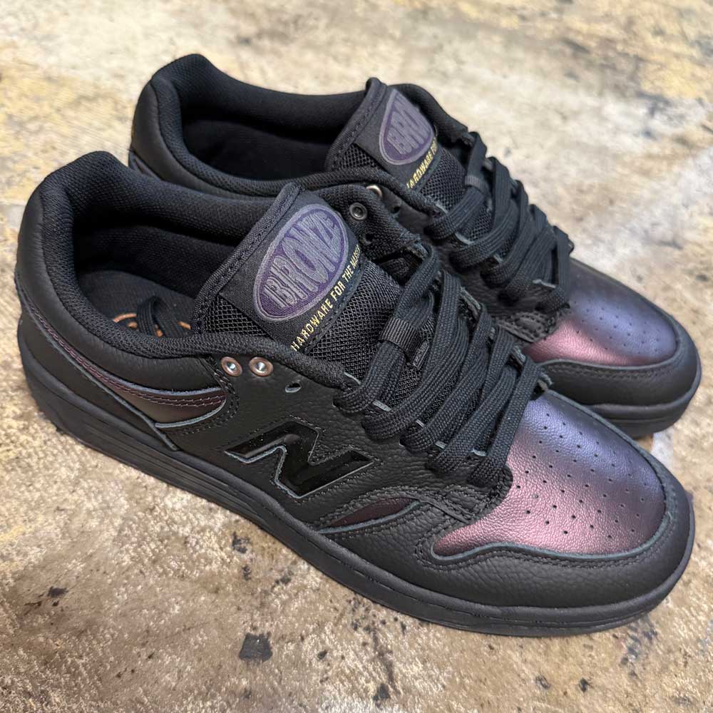 A pair of 480 BFK Bronze 56K New Balance Numeric Skateboarding Shoes in suede black iridescent with a durable leather upper, black laces, and tongue branding, displayed on a textured beige surface.