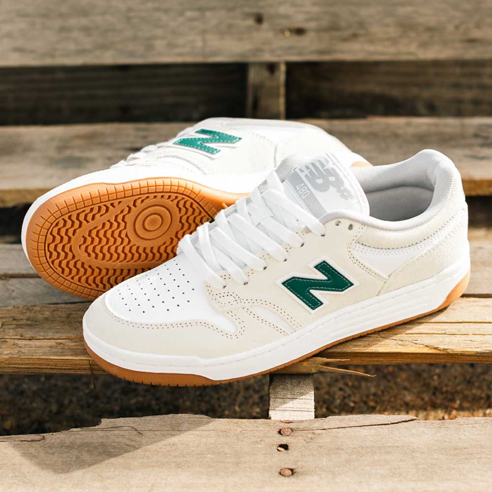 A pair of 480 JDI New Balance Numeric Shoes in Suede White Green, featuring green N logos and gum soles, are shown on wooden steps—one upright and one on its side to highlight the durable sole.