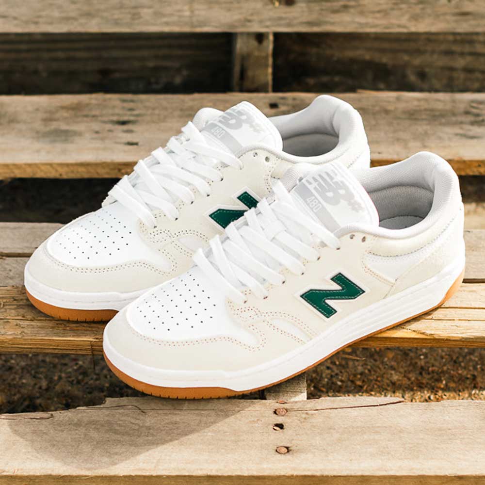 A pair of 480 JDI New Balance Numeric Shoes in suede white with green accents and gum soles sits on wooden steps under natural light.