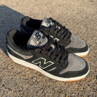 A pair of 480 MGC New Balance Numeric Shoes in Suede Black and Dark Grey with white details and gum soles are set on a textured concrete surface under bright sunlight.