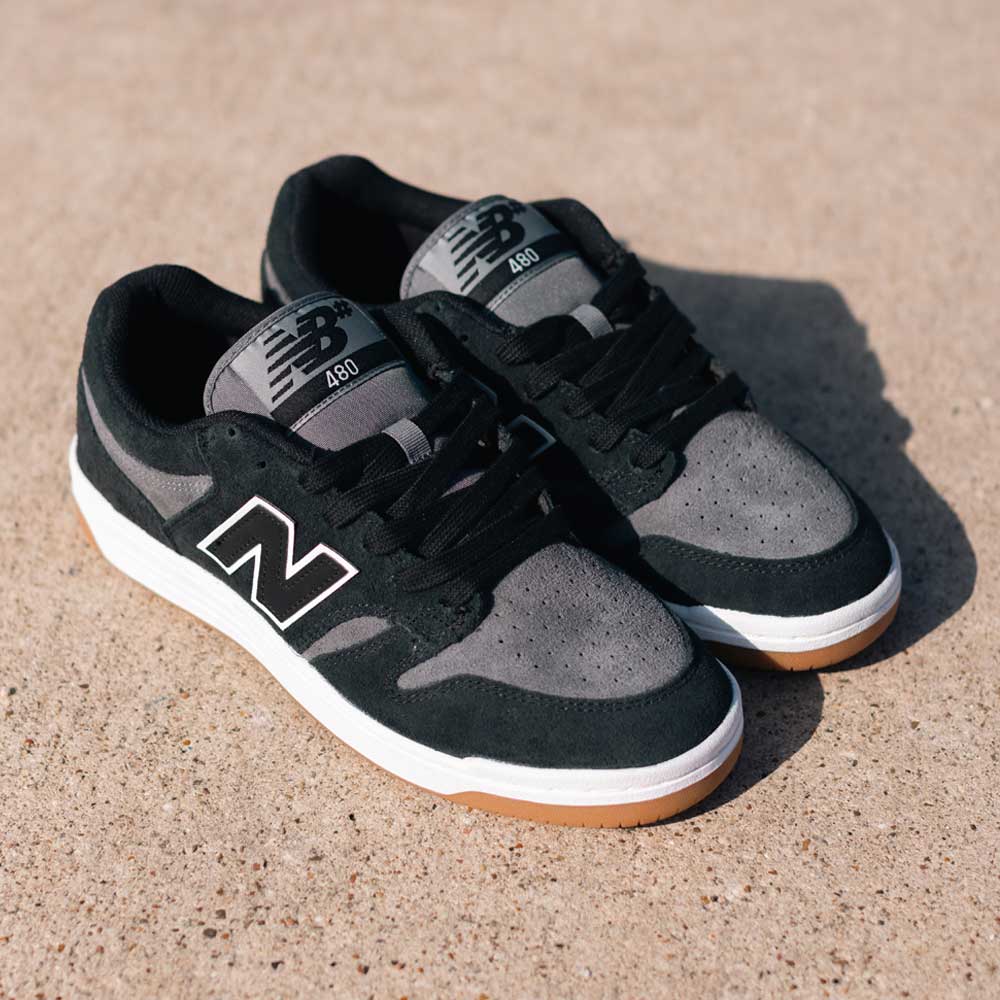 A pair of 480 MGC New Balance Numeric Shoes in suede black and dark grey with white soles and gum bottoms sits on a sunlit concrete surface.