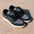 A pair of 480 MGC New Balance Numeric Shoes in suede black and dark grey with white soles and gum bottoms sits on a sunlit concrete surface.