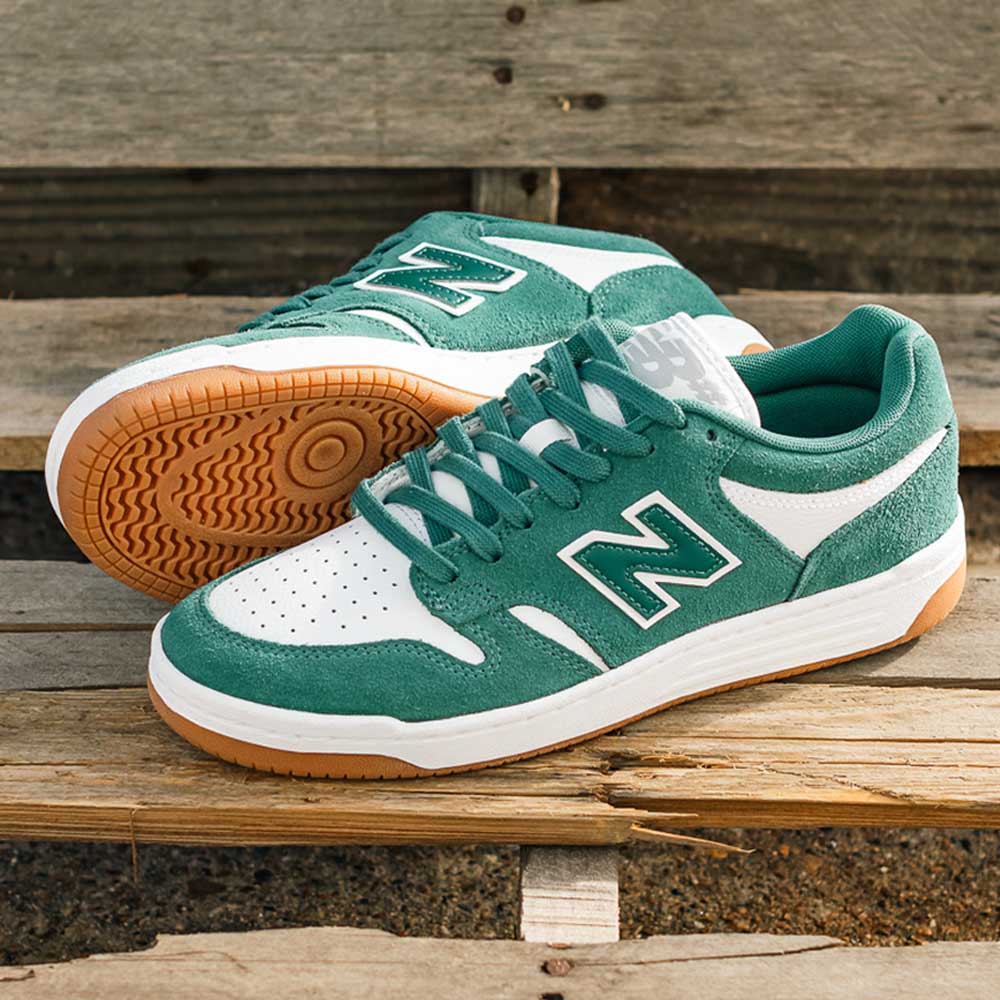 A pair of 480 NDI New Balance Numeric Skateboarding Shoes in suede green and white with gum soles and a durable upper are displayed outdoors on wooden benches, one upright and the other on its side.