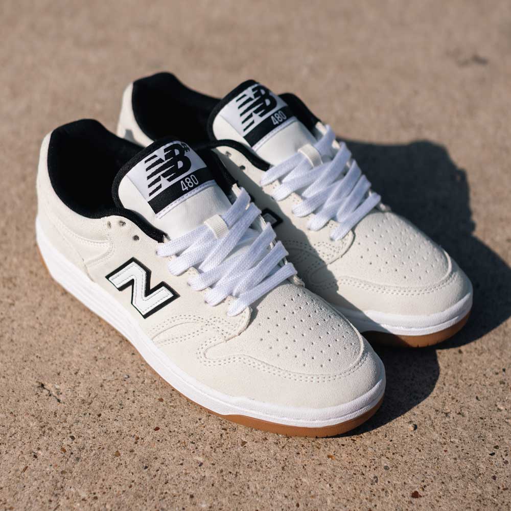 A pair of 480 SWG New Balance Numeric Shoes in suede white and black with gum soles are shown on a sunlit concrete surface.