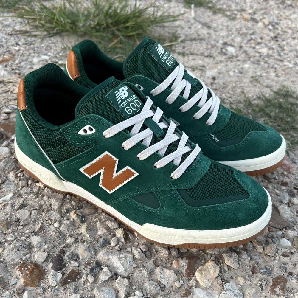 A pair of 600 ALB Tom Knox Green Brown Suede New Balance Numeric Skateboarding Shoes with white laces and soles are placed on gravel, with patches of grass visible in the background.