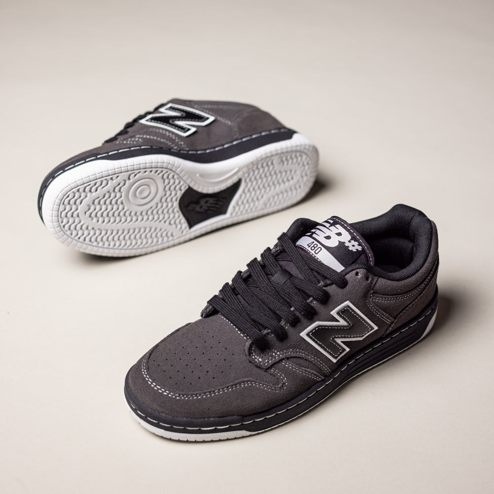 A pair of Black Cement 480 CSS New Balance Numeric Suede Skateboarding Shoes with white soles—one upright showing the side and top, the other flipped to display the sole—rest on a light, neutral background. Inspired by heritage basketball styles.