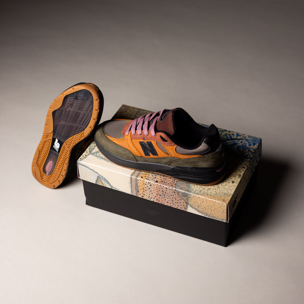 The Mike Gigliotti True Brown Candy Pink 933 LOT New Balance Numeric Suede Skateboarding Shoes, featuring brown, orange, and black accents, are displayed—one on a patterned box, the other on its side to reveal the sole. The neutral background is softly lit.