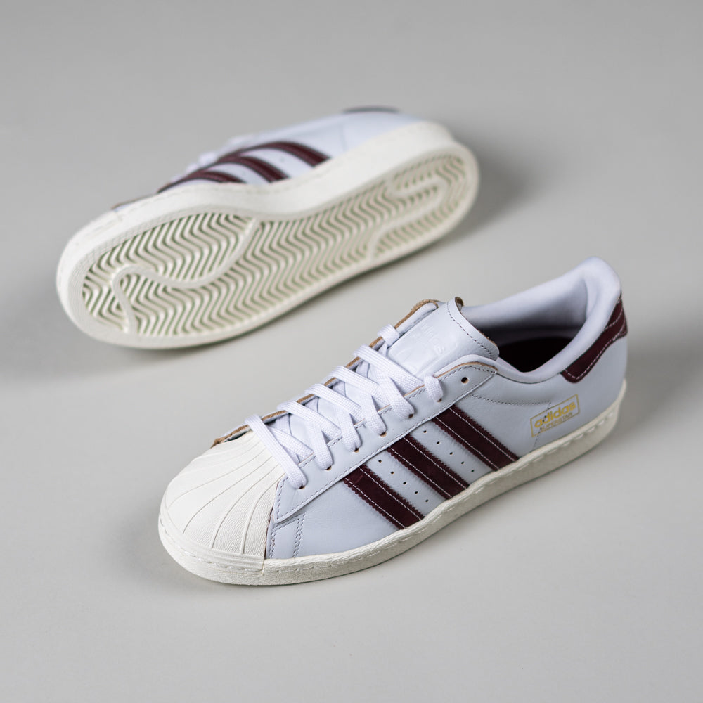 A pair of JP8529 Cloud White Share Superstar 80 ADV Adidas Skateboarding leather shoes with dark burgundy stripes and gold detailing are displayed on a light gray surface; one shoe is upright, the other shows the sole.