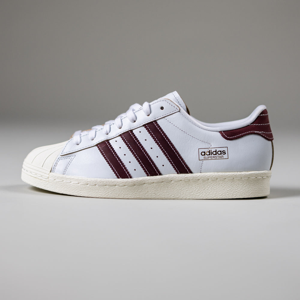 The JP8529 Cloud White Share Superstar 80 ADV Adidas Skateboarding leather shoes feature a white upper with maroon stripes, cream rubber sole, and classic shell toe—ideal for stylish skateboarding—shown on a plain grey background.