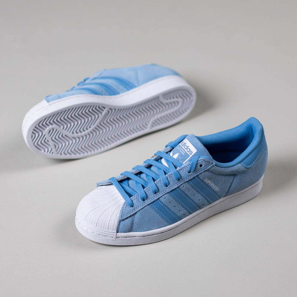 A pair of JQ1102 Light Blue White Superstar ADV Adidas Skateboarding suede shoes, featuring blue stripes and laces, are displayed on a light gray surface—one upright, the other on its side revealing the sole.