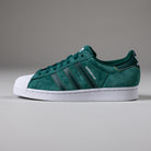 The JQ1103 Core Green White Superstar ADV Adidas Skateboarding Suede Shoes, featuring black stripes, a white shell toe, and "SUPERSTAR" in white on the side, are shown on a plain light grey background.