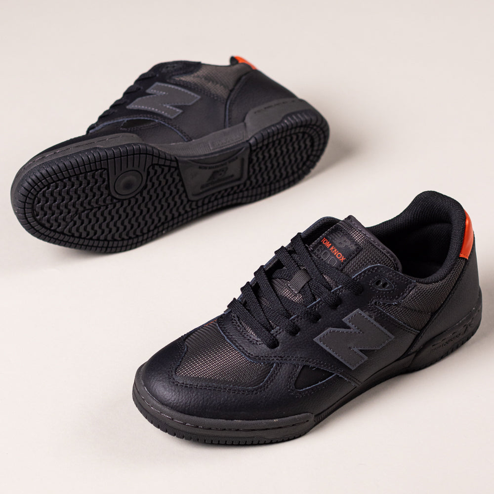 A pair of 600TOO Black Orange Tom Knox New Balance Numeric Leather Skateboarding Shoes are displayed on a light background, with one upright to reveal the side and top, and the other on its side showing the sole and FuelCell cushioning.