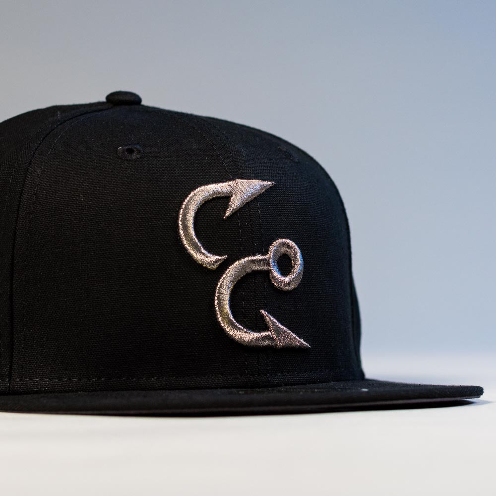 The Eagle Claw Canvas Black Corpus Christi Hooks New Era Hat 5950 Fitted is a black cap with a flat brim and a silver embroidered design of two fish hooks forming a stylized C on the front. The softly lit background is plain.