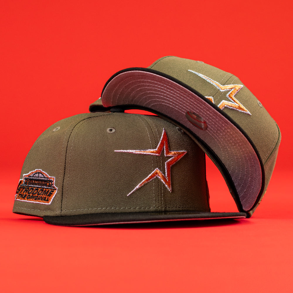 Two The Original Olive Black CC Astros New Era Hat 5950 Fitted caps with orange and white star logos, black visors, and commemorative patches are displayed on a bright red background—one upright, one tilted to show the underside.
