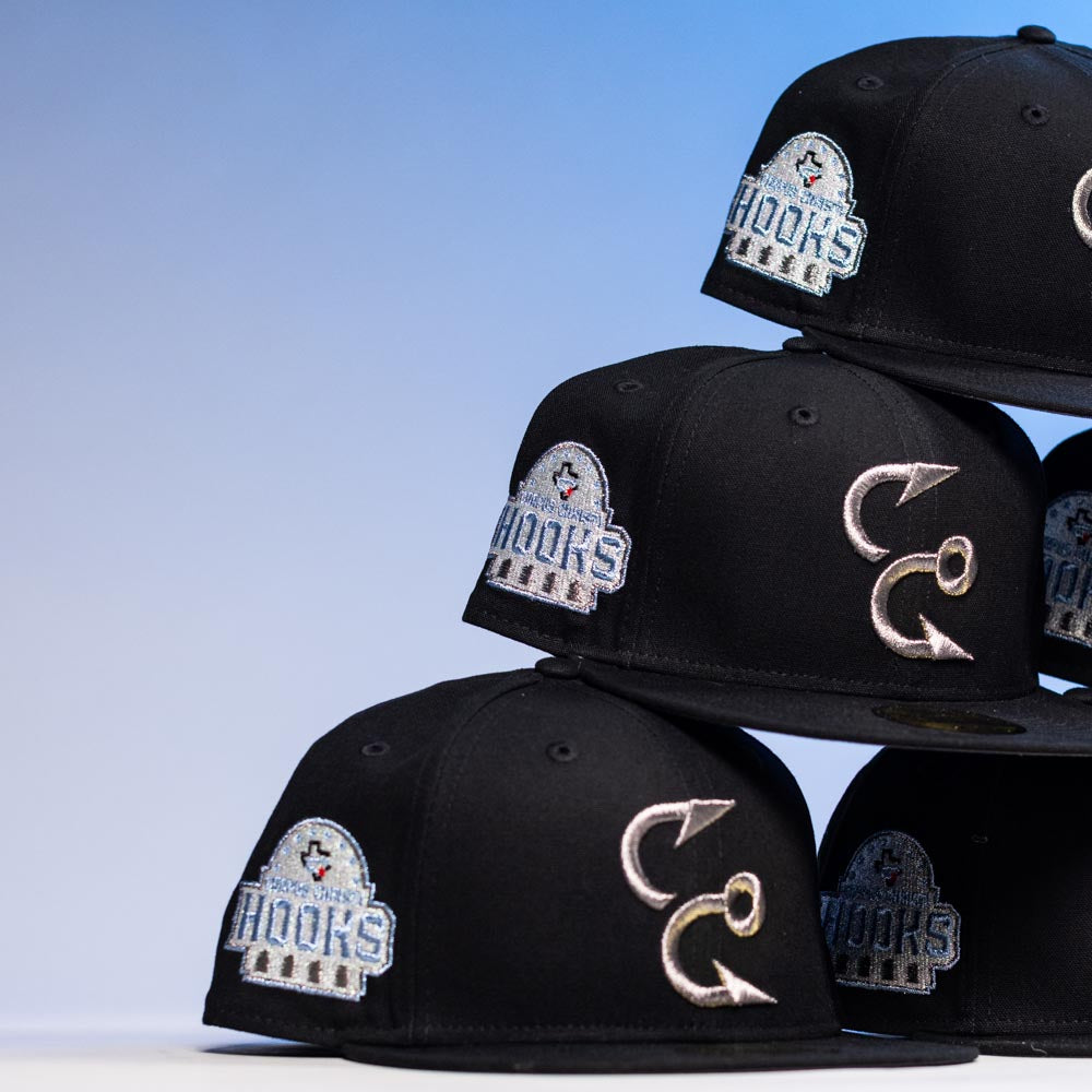 The Eagle Claw Canvas Black Corpus Christi Hooks New Era 5950 Fitted Hat features embroidered team logos, a fishing hook, and a fish emblem. Displayed in a stack against a gradient blue background.