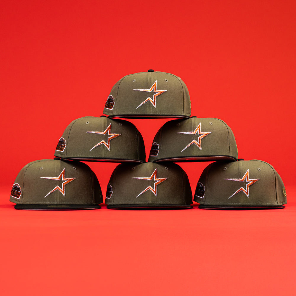 Six The Original Olive Black CC Astros New Era Hat 5950 Fitted caps, featuring the orange Astros star logo, are stacked in a pyramid against a bright red backdrop.