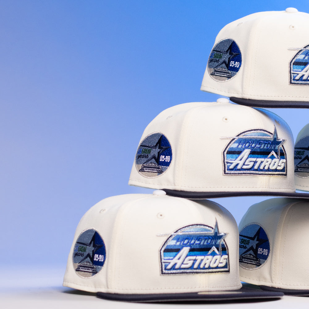 A stack of KB1 Rushmore 35th Chrome Navy Houston Astros New Era Hat 5950 Fitted caps with blue brims and team patches are arranged against a blue gradient background.