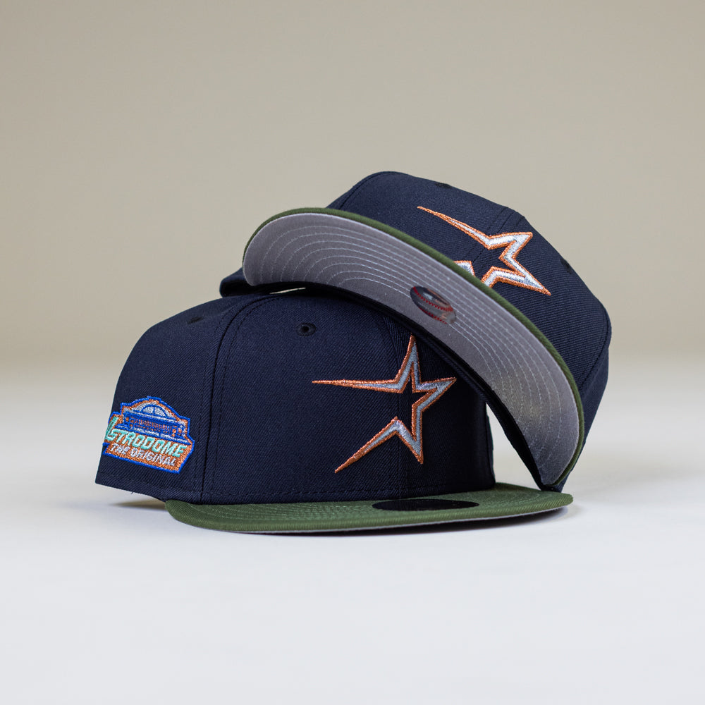 Two Aquamarine Navy Rifle Green Astros New Era Hat 5950 Fitted caps, each with an orange and white star logo—one upright showing the “Astrodome Commemorative” patch, the other resting on its side.