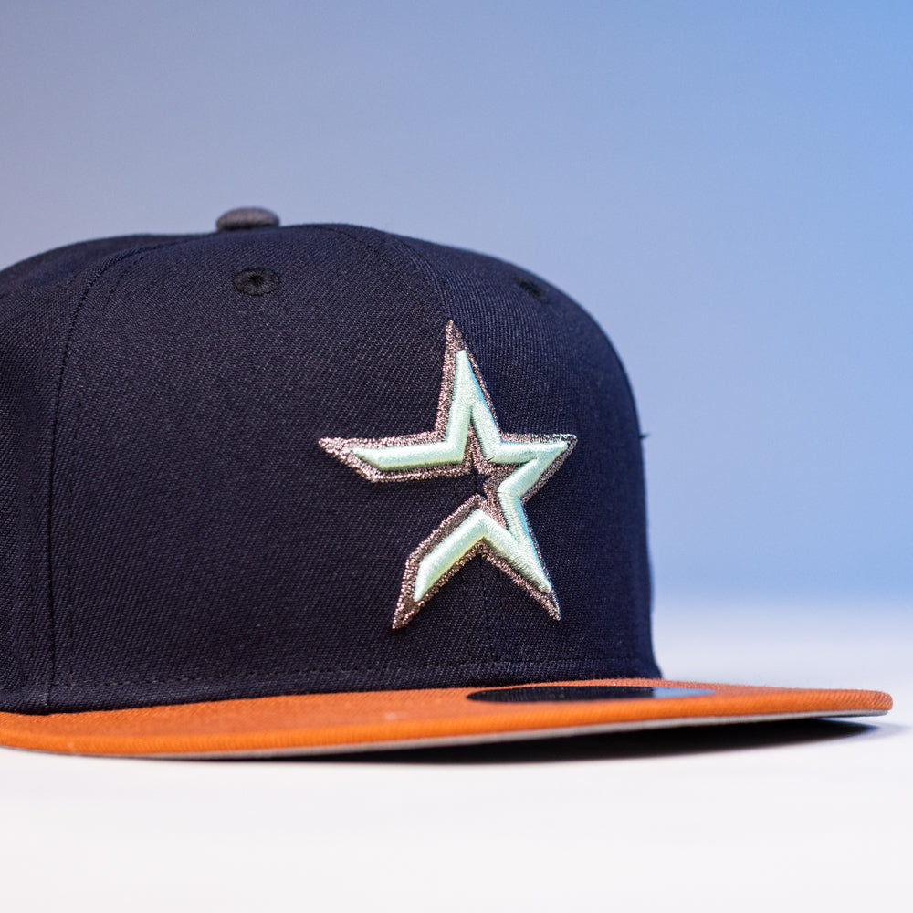 The Feast Navy Rust Houston Astros New Era Hat 5950 Fitted features a stylized silver and green star logo on the front and an orange brim, shown against a light blue background—perfect for Astros fans.