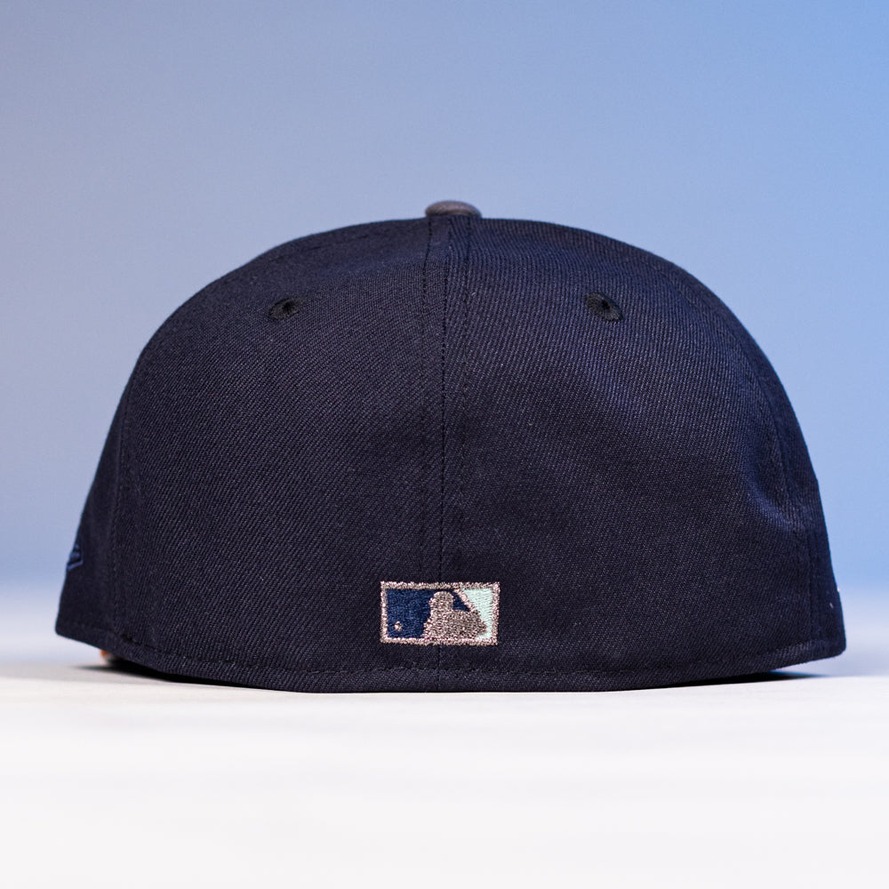 The Feast Navy Rust Houston Astros New Era 5950 Fitted Hat, shown from the back, displays a small embroidered MLB logo near the bottom center against a softly lit blue and white background.