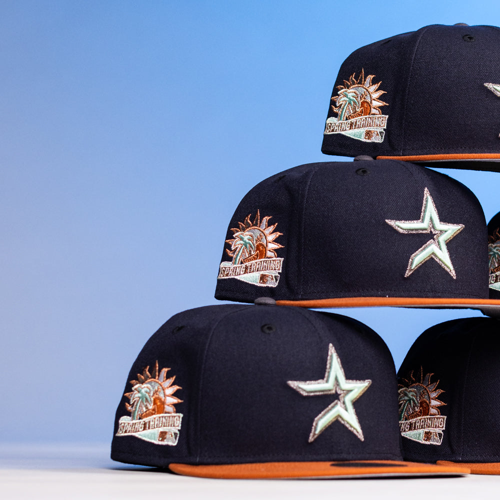 Six The Feast Navy Rust Houston Astros New Era Hat 5950 Fitted caps are stacked in two piles. Each features a star emblem on the front and "Spring Training" palm tree side patches. The hats have orange brims; background is light blue.