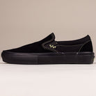 The VN000EKPGSH Black Black Olive Skate Slip On Vans Suede Shoe features a black suede upper, black sole, light green stitching, and a small checkerboard accent by the side elastic. It's shown in profile on a beige background.