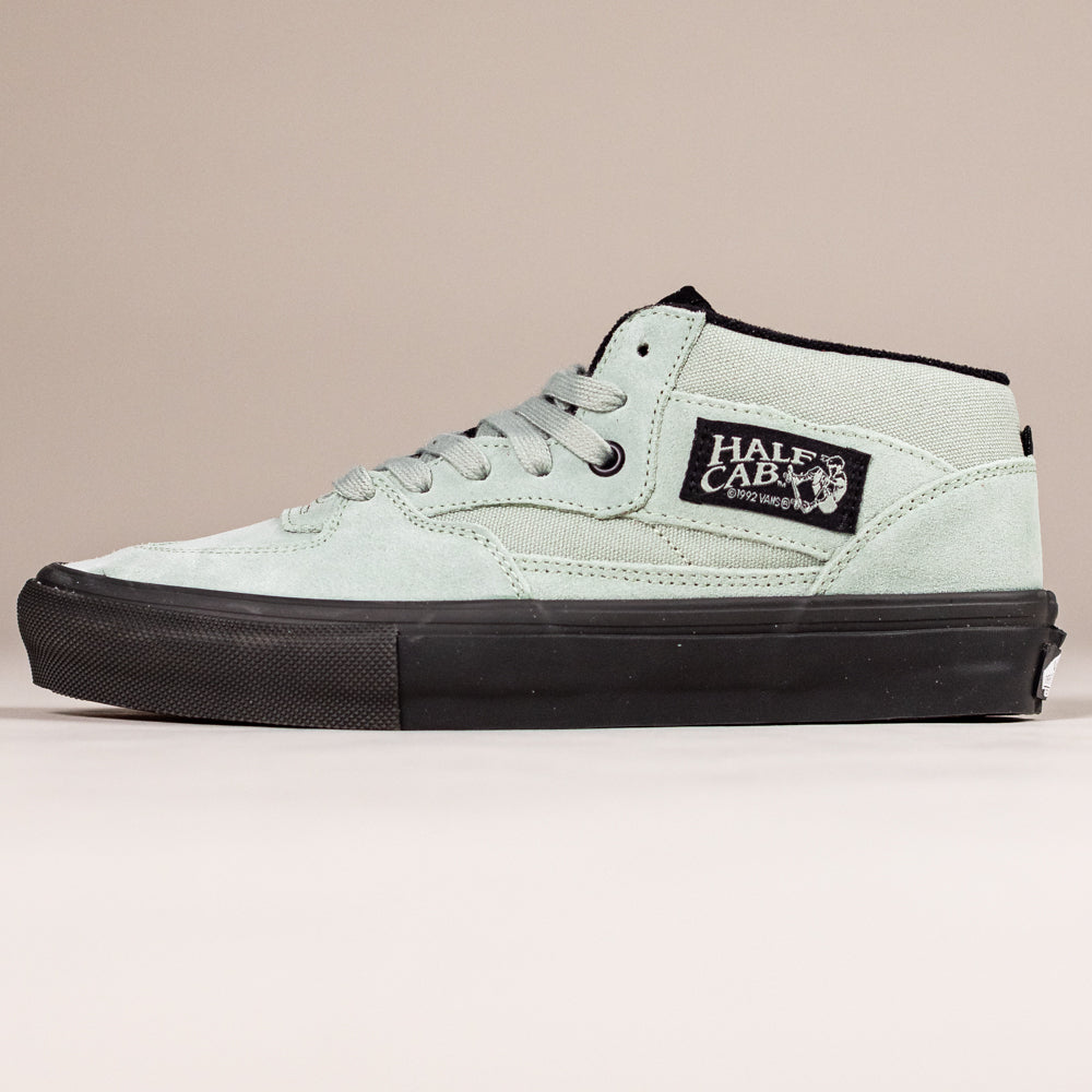 The VN0A5FCDEMW1 Gray Olive Suede Skate Half Cab Vans is a mid-top skate shoe with black laces, black rubber sole, black HALF CAB patch with white text, PopCush cushioning, displayed on a plain beige background.