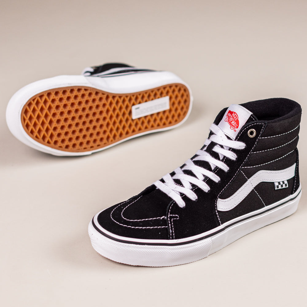 A pair of VN0A5FCCY281 Black White Skate SK8-HI Suede Vans shoes: high-tops with white laces, white soles, and a wavy stripe. One stands upright; the other lies on its side revealing the brown waffle-patterned sole.