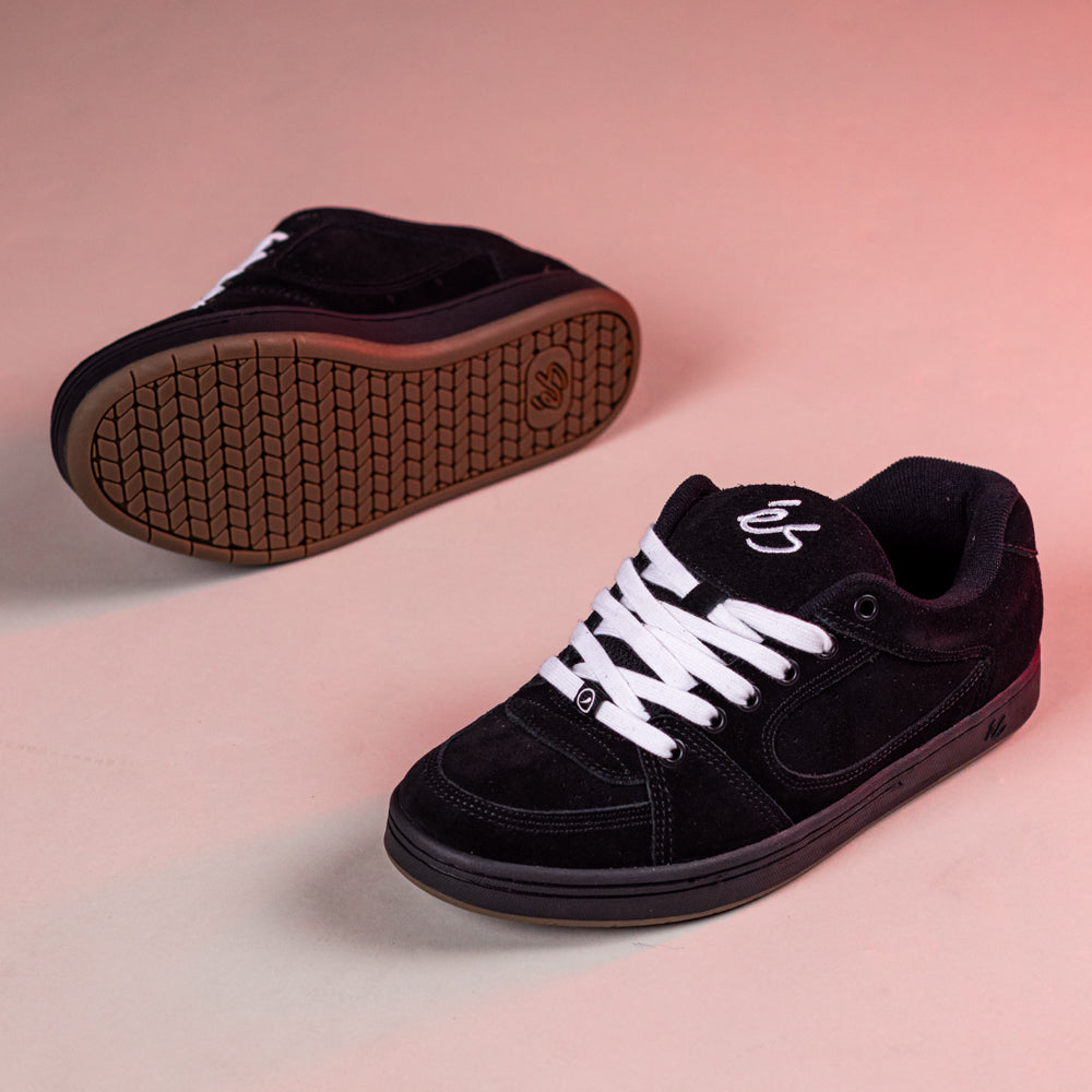 A pair of Accel Black Noir Es Shoes Skateboarding Original Shape with white laces and a white side logo are shown on a light pinkish surface; one is upright, the other displays the brown cupsole, reflecting classic 1995 skate style.