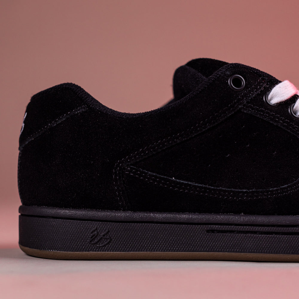 A close-up of the Accel Black Noir Es Shoes shows their black suede upper, black laces, and detailed stitching—skateboarding originals inspired by the 1995 design—set against a pinkish background with the side and rear visible.