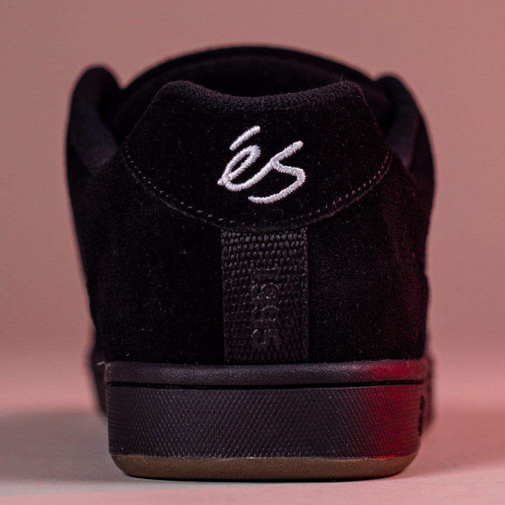 Close-up rear view of the Accel Black Noir Es Shoes Skateboarding Original Shape, featuring black suede, white embroidered éS branding on the heel tab, and "Sole" text on the heel strap, set against a pinkish background.