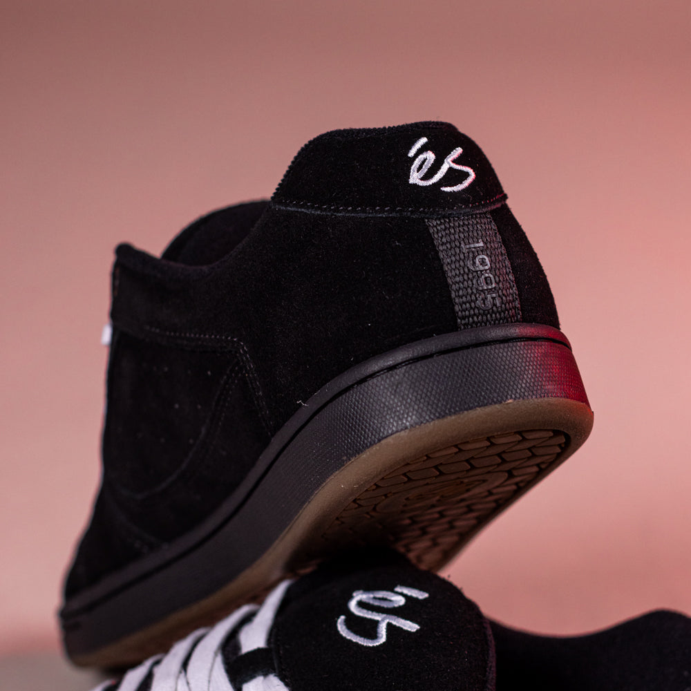 Close-up of Accel Black Noir Es Shoes Skateboarding Original Shape in black suede, showing the white “es” logo on heel and tongue with a gum rubber outsole. These classic cupsole shoes are stacked against a pink background.