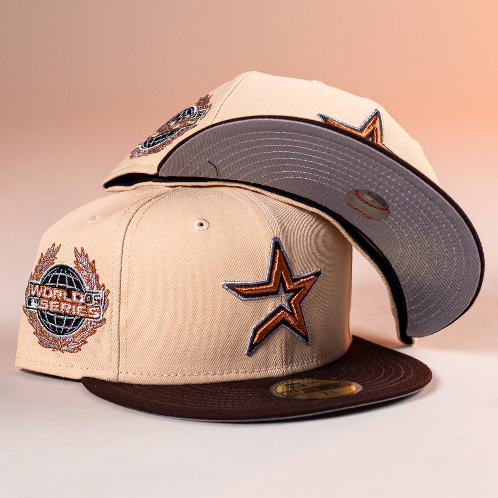 Two Flat White WS05 Mango Wood Astros New Era Hat 5950 Fitted caps are displayed. One lies flat showing a star logo and World Series patch; the other is balanced on top, revealing an embroidered baseball under the brim.