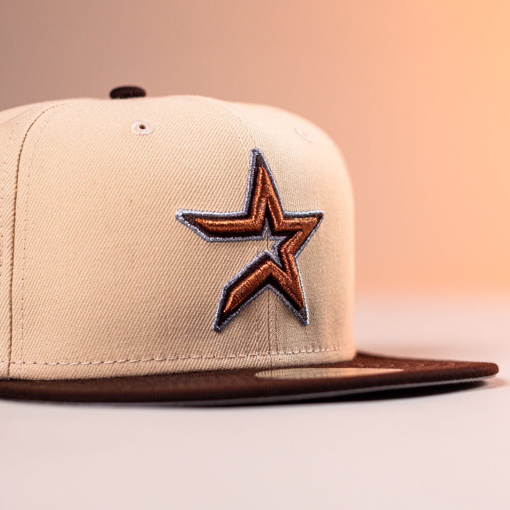 The Flat White WS05 Mango Wood Astros New Era Hat 5950 Fitted, with a beige crown, brown brim, and orange/black outlined star logo on the front, is shown against a softly lit peach background.