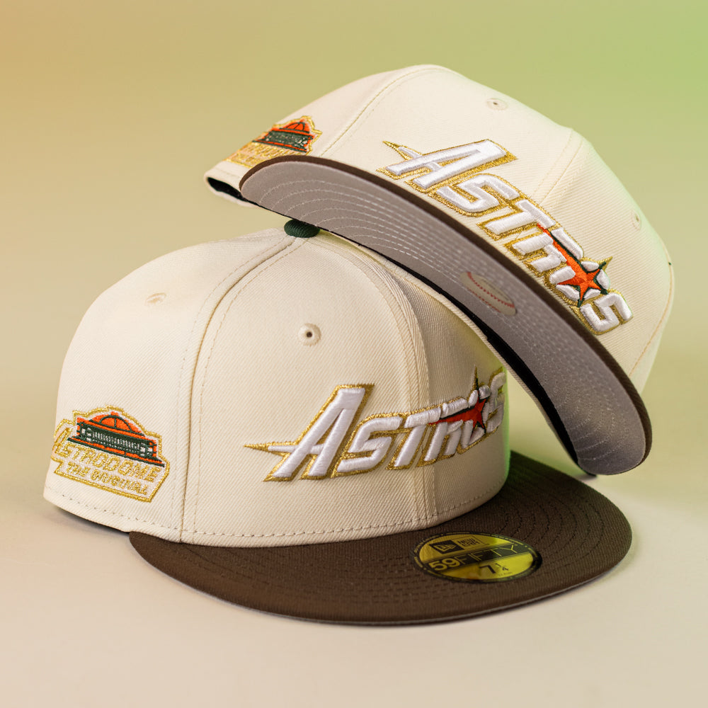 Two Oompa Pink Box Houston Astros 5950 Fitted New Era Hats are stylishly displayed, featuring cream color, brown brims, embroidered Astros logos, and a colorful side patch—one cap upright and the other leaning on top.
