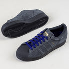 The JR7409 Carbon Purple Superstar ADV Adidas suede skateboarding shoes feature textured dark grey uppers, black shell toes and soles, blue laces, and gold branding. Displayed on a light background, one shoe stands upright, the other is on its side.