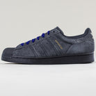 The JR7409 Carbon Purple Superstar ADV Adidas Suede Skateboarding Shoes feature a dark gray, carbon purple suede upper, black rubber sole and toe cap, blue laces, and gold SUPERSTAR branding—shown on a light gray background.