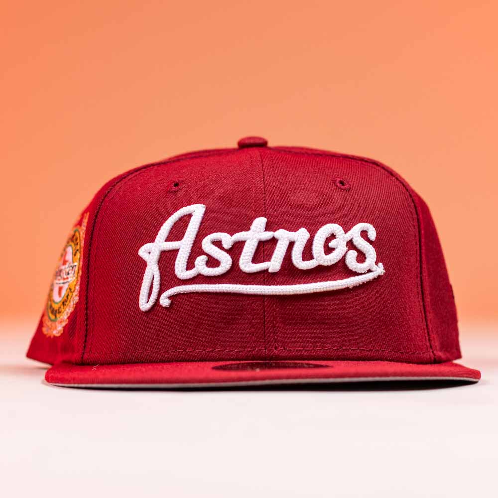 The Big Puma Cardinal White Script New Era Hat 5950 Fitted, featuring Astros in Cardinal White script on the front, is shown in red against an orange background.