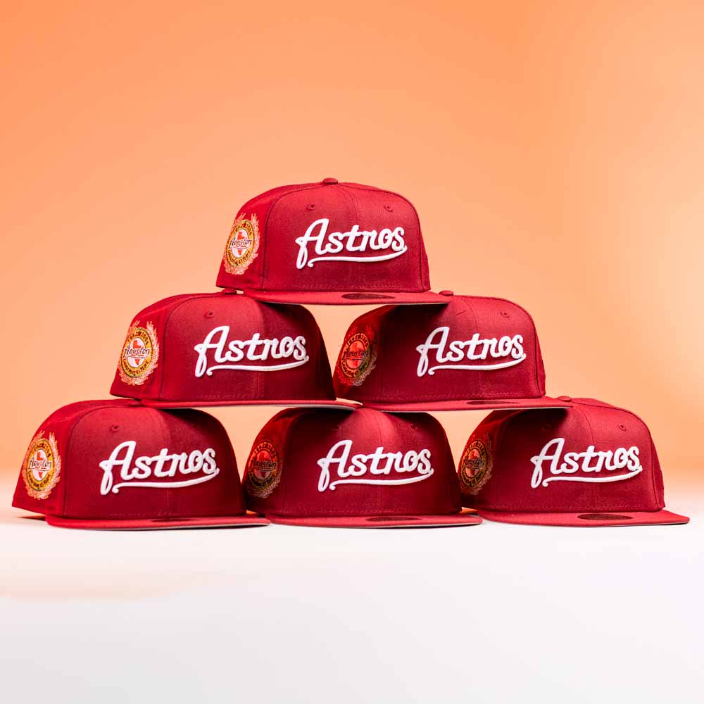 Six red The Big Puma Cardinal White Script New Era Hat 5950 Fitted caps are stacked in a pyramid against an orange background, each featuring the Astros logo on the front, a circular side patch, and white script details.