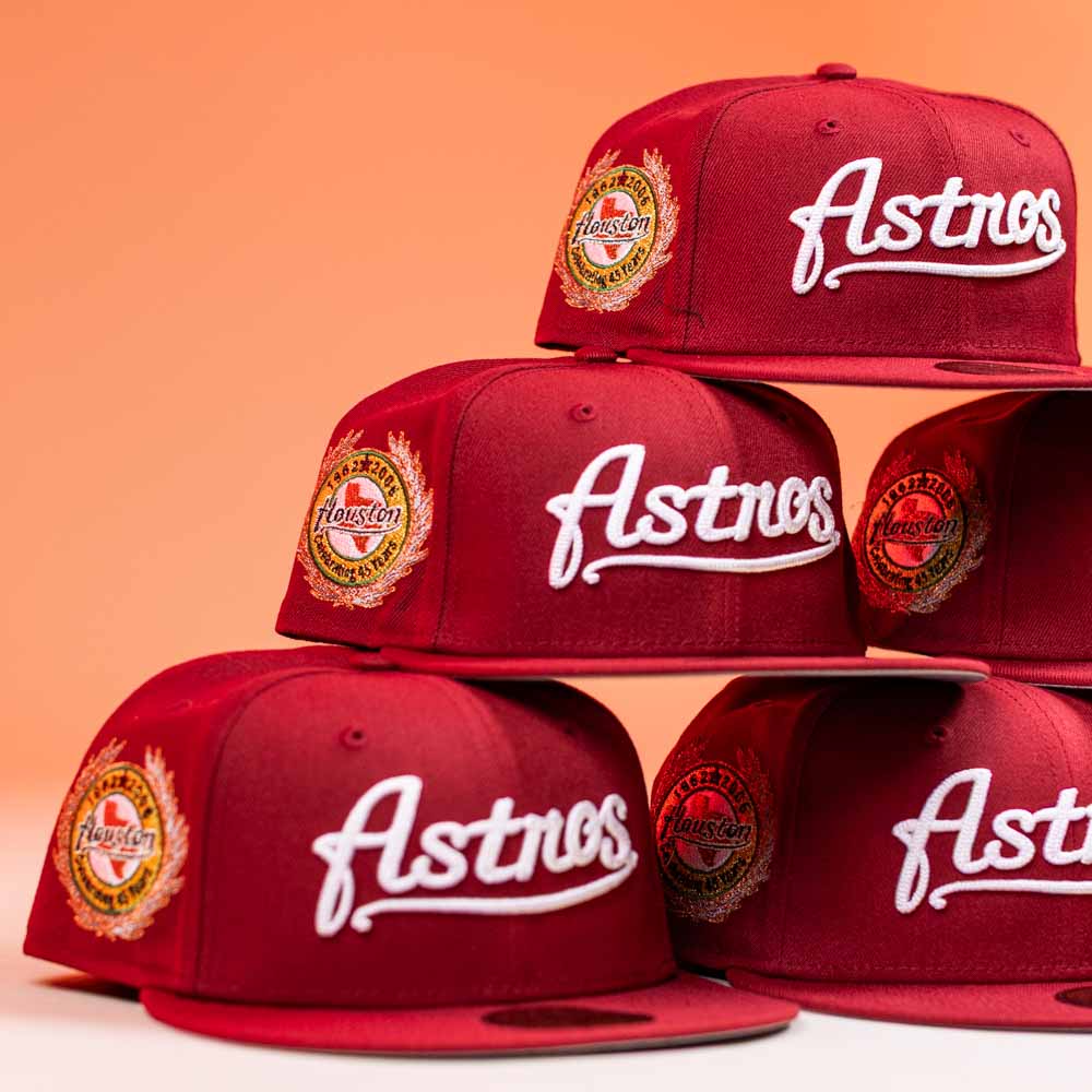 Several The Big Puma Cardinal White Script New Era Hat 5950 Fitted hats in red are stacked, each with bold "Astros" script and a Houston Astros patch. The orange-white gradient background highlights these standout styles.