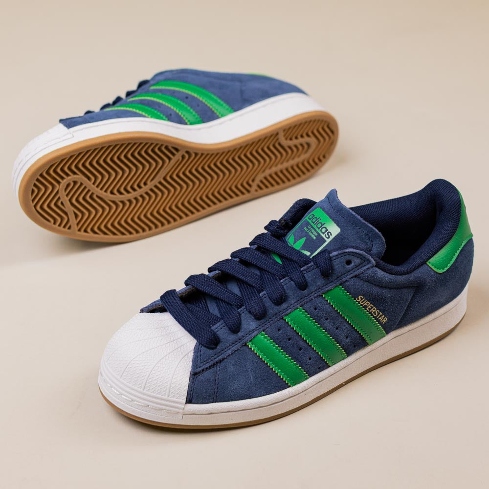 The JQ2098 Navy Green White Superstar ADV Adidas Suede Skateboarding Shoes feature green stripes, white shell toes, tan cupsoles, and durable construction, displayed on a light beige background.