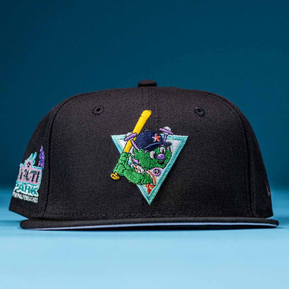 The Flamingo and Chill Bo Bundy 2022 Houston Astros New Era Hat 5950 Fitted is black with a colorful embroidered mascot holding a yellow bat, wearing a helmet, and standing before a teal triangle. It’s shown on a blue surface with a blue background.